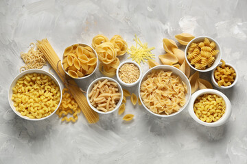 Bowls with different types of raw pasta on grey grunge background