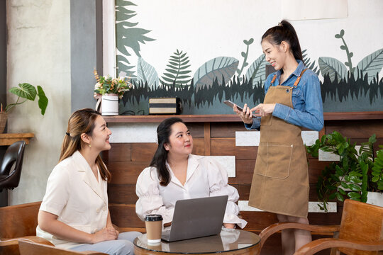Young female barista smiling while taking an order from two women seated with coffee and a laptop, capturing a cheerful moment of service in a welcoming local coffee shop.