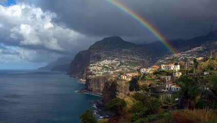 A scenic coastal view of mountainous terrain beneath dramatic clouds and a vibrant rainbow