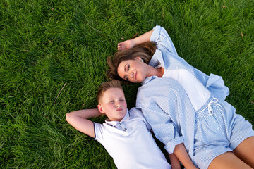 Overhead view of a relaxed mother and her son lying on thick green grass in a summer park, sharing...