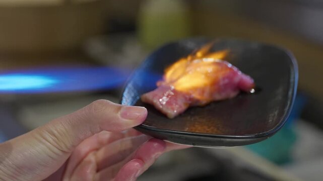 A seared beef aburi sushi being blow torched with a flame on a black plate in a Japanese restaurant