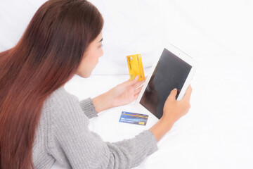 Over-the-shoulder view of woman holding credit card and tablet indicating online browse or budgeting for shopping on white bed.