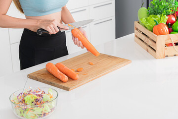 Close-up woman in athletic wear is preparing hand cutting fresh carrot on cutting board with other vegetable and salad in the background.