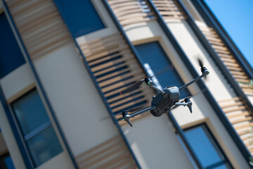 A drone hovers in mid-air, providing an aerial perspective of a contemporary building with striking wooden accents against a bright sky, showcasing its design and urban environment.