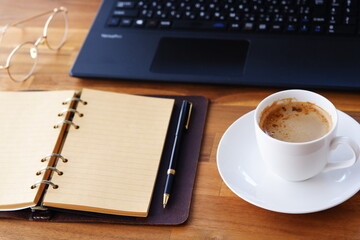 机にリング式のシステム手帳を広げ、淹れたてのコーヒー（カフェ・ラテ）を飲みながら、パソコンでデスクワークをしているイメージ
