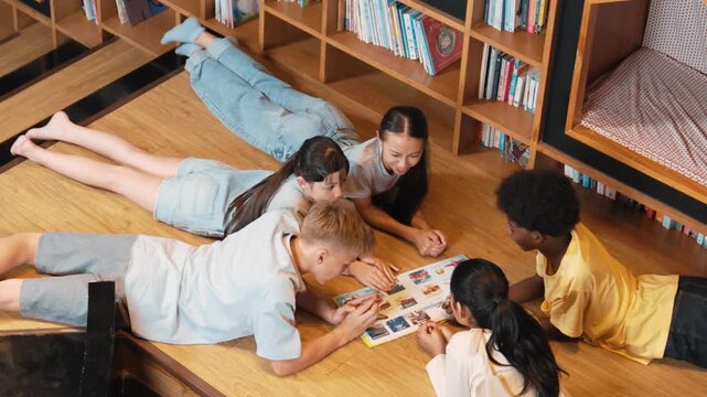 Group of diverse children lying down in circle while reading a book at library. Top view of girl sharing a magazine while pointing at interested topic and talking with lovely friends. Edification