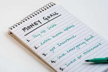 Spiral notebook with money goals list written in teal ink and green pen on white background for...