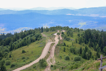 A mountain landscape with a dirt road winding along green slopes against the backdrop of blurred silhouettes of distant mountains. The bright part of the day emphasizes the natural beauty and invites 