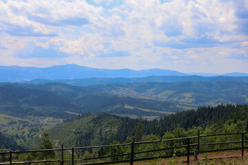 A panoramic view of majestic mountain ranges that smoothly transition into green valleys filled with forests and scattered houses.