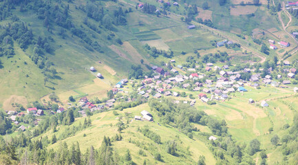 An aerial view of a picturesque mountain village surrounded by green hills and forests. Residential houses with colorful roofs are scattered among well-kept fields and meadows, creating an atmosphere 