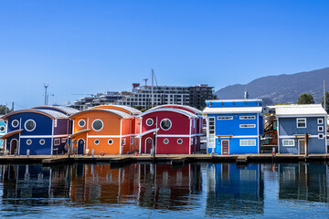 Row of vividly painted floating homes moored at a North Vancouver marina: circular porthole windows, bold primary colours and Canadian flags mirror crisply in the still harbour water beneath.