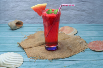 Refreshing summer drink with watermelon and ice in a glass, decorated with fruit slices, mint, and a straw, on a rustic blue wooden table with tropical details