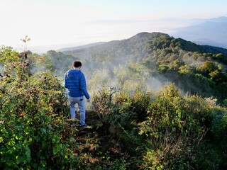 Naklejka premium Person in blue jacket overlooking misty hills at Doi Pha Hom Pok National Park, Thailand. Serene mountain landscape with lush greenery and tranquil atmosphere.