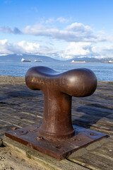 Rusty mooring bollard on Vancouver’s weathered pier overlooks English Bay, cargo ships and coastal mountains beneath a bright summer sky.