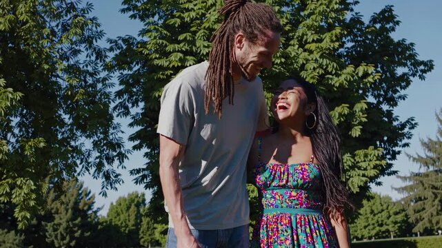 A candid video still of a smiling couple outdoors, shot from a low angle. The bright, sunny day highlights their casual, colorful attire and natural setting.