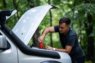 Driver connecting jumper cables to car battery in forest