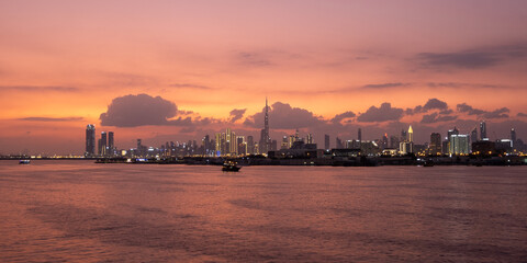 Panoramic view of Dubai skyline from Dubai Creek Harbour waterfront at sunset, modern skyscrapers and iconic Burj Khalifa reflecting on calm water