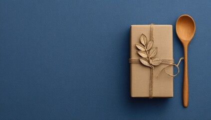 Gift box with natural wrapping, tied with twine, and a decorative leaf, next to a wooden spoon, on a deep blue background
