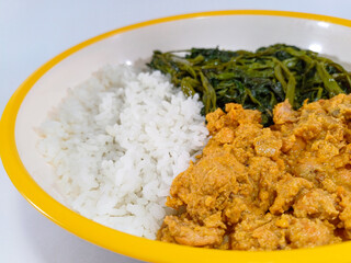 Plate of food containing white rice, a golden-brown curry or stew, and stir-fried green vegetables againts a white background. Related to Southeast Asian cuisine, home-cooked meals, or food and drink