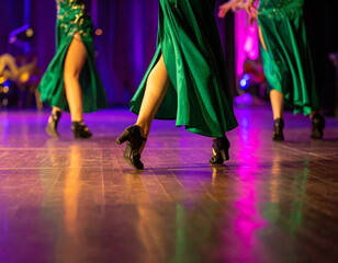 Low angle shot of dancers in long green dresses with high slits and black heels on a shiny wooden stage with vibrant lighting.