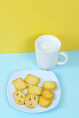 A plate of various shortbread cookies and a white mug of milk sit on a blue surface against a yellow wall.