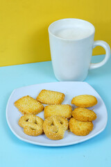 A plate of various shortbread cookies and a white mug of milk sit on a blue surface against a yellow wall.