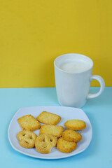 A plate of various shortbread cookies and a white mug of milk sit on a blue surface against a yellow wall.