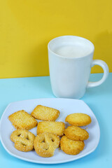 A plate of various shortbread cookies and a white mug of milk sit on a blue surface against a yellow wall.