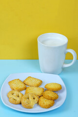 A plate of various shortbread cookies and a white mug of milk sit on a blue surface against a yellow wall.