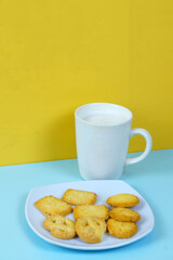 A plate of various shortbread cookies and a white mug of milk sit on a blue surface against a yellow wall.