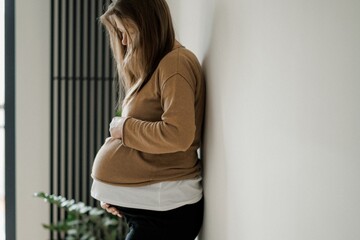 A pregnant woman gently cradles her baby bump while standing against a neutral background. She wears a cozy brown sweater and looks down with a serene expression.