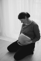 A pregnant woman gently cradles her baby bump while standing against a neutral background. She wears a cozy brown sweater and looks down with a serene expression.