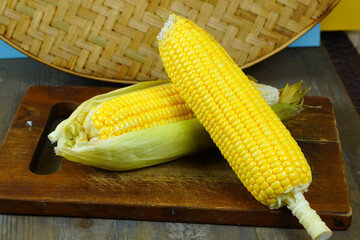 Two fresh, yellow corns, one partially husked, rest on a rustic wooden cutting board with a woven basket in the background.