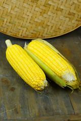 Two fresh, yellow corns, one partially husked, rest on a rustic wooden cutting board with a woven basket in the background.