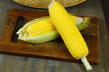 Two fresh, yellow corns, one partially husked, rest on a rustic wooden cutting board with a woven basket in the background.
