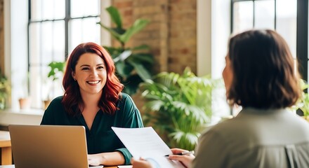 Two women in a modern office discussing documents with a laptop present