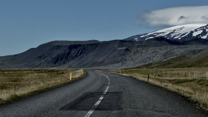 Empty road leading through the remote Icelandic countryside with volcanic cliffs and glacier in the background, under a clear sky.