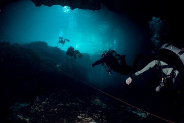 Divers explore a dark underwater cave system, illuminated by a beam of light.