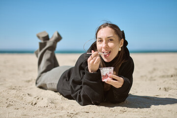 Young woman with long brunette hair savoring yogurt on sandy beach. Bright, sunny day enhances...