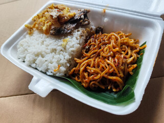Styrofoam box containing mixed rice, with white rice, fried salted fish, and fried noodles covered in banana leaves, ready to eat.