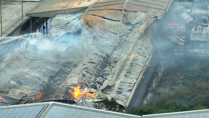 Aerial drone view of firefighters spraying water on a massive industrial plant fire, battling...