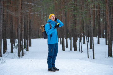 Hiker wearing a blue jacket and yellow beanie searching for cellphone signal in a snowy forest, surrounded by tall trees and winter's chill during a solitary adventure