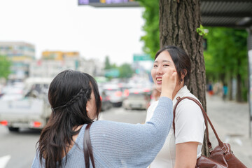On a bustling boulevard, two Korean women in their twenties in casual clothes stand at a bus stop,...