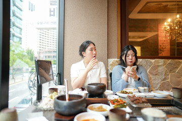 In a kalbi barbecue restaurant at a sunlit window table, two Korean women in their twenties in...