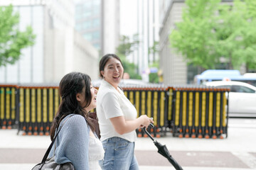 Two Korean women in their twenties in casual clothes walk along a boulevard sidewalk, talking and...