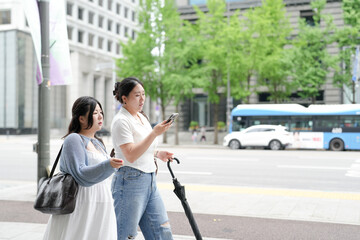 Two Korean women in their twenties in casual clothes walk along a boulevard sidewalk, talking and...
