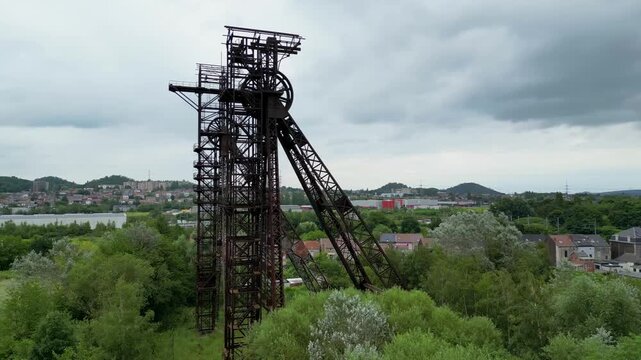 Aerial view of the imposing rusty, steel structures of Bois du Cazier rise amidst the lush greenery, contrasting against a backdrop of the urban landscape, Charleroi, Hainaut, Belgium.