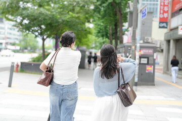 Two Korean women in their twenties in casual clothes walk along a boulevard sidewalk, talking and...