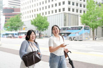 Two Korean women in their twenties in casual clothes walk along a boulevard sidewalk, talking and...