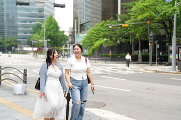 Two Korean women in their twenties in casual clothes walk along a boulevard sidewalk, talking and...